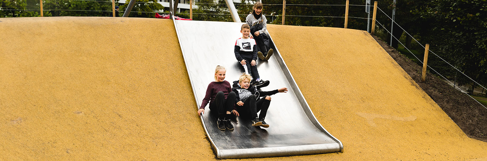 Children slide down a wide embankment slide with smiles on their face at a playground.